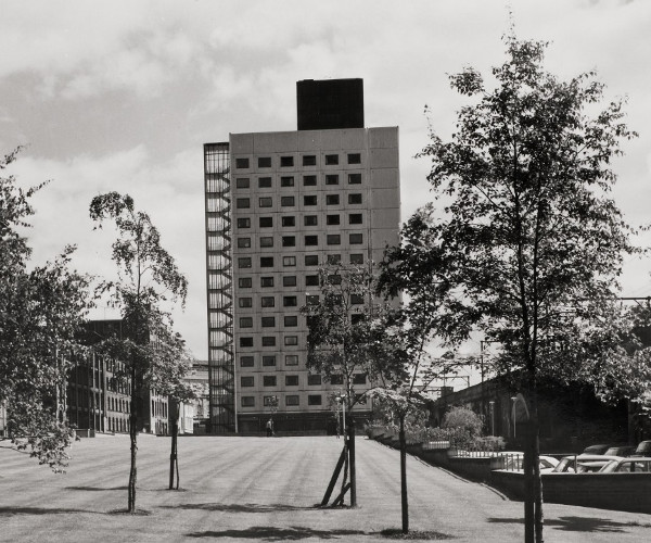 View across a green of Chandos Hall, a 1960s tower block, framed by the Main UMIST Building.