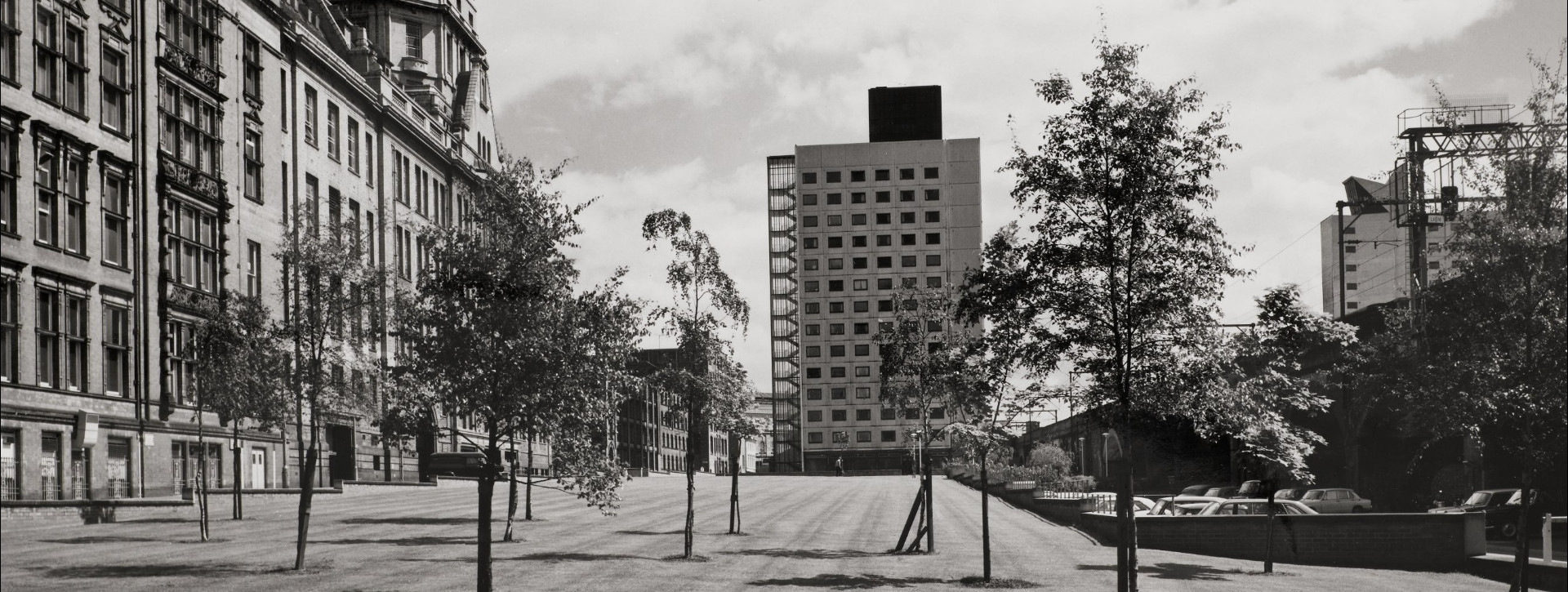 View across a green of Chandos Hall, a 1960s tower block, framed by the Main UMIST Building.