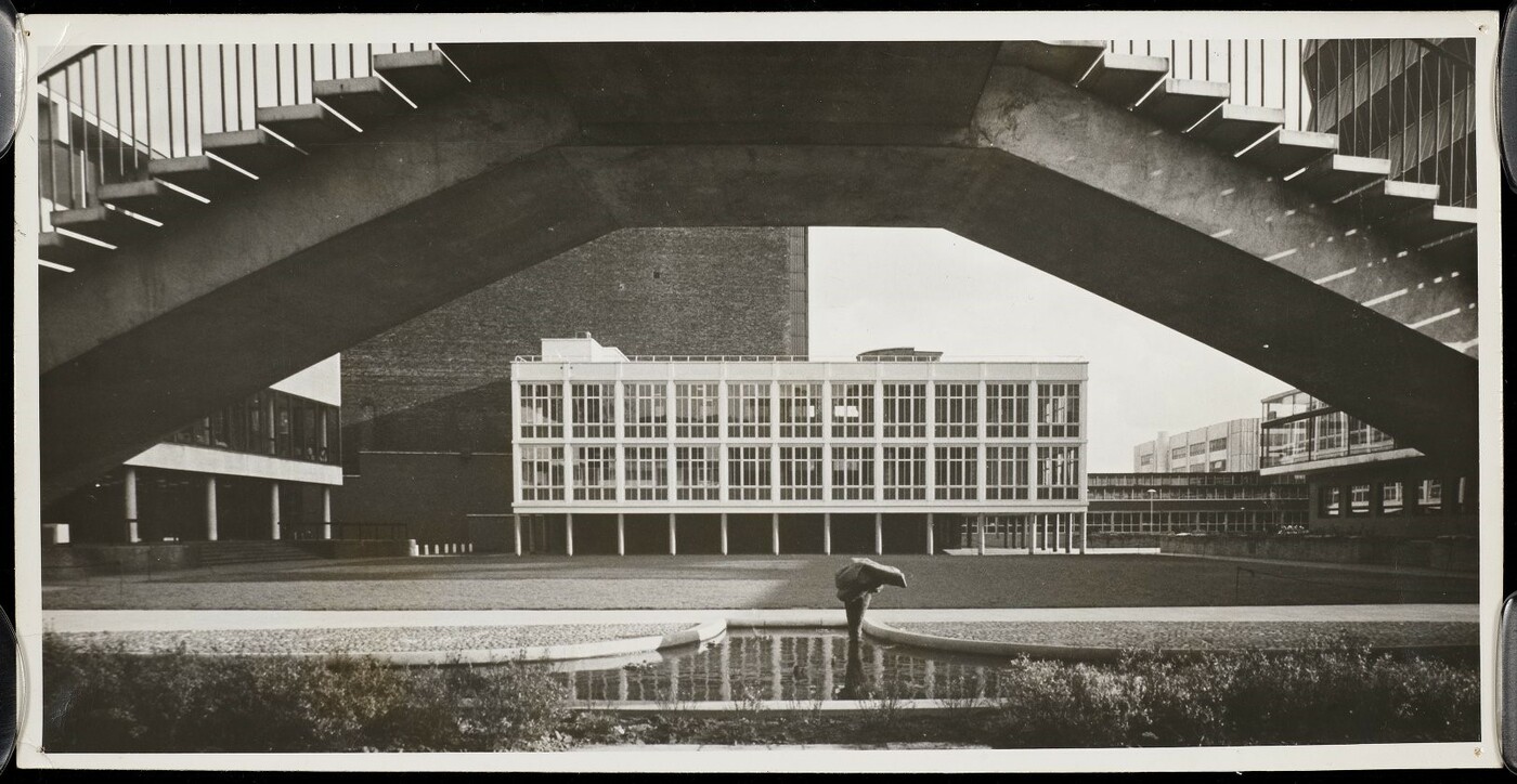 View of a modernist glass building in the medium distance, framed by an elevated concrete pedestrian bridge.