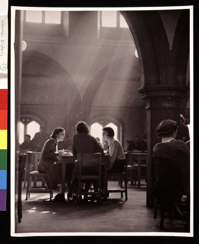 Photograph of interior of the students union coffee bar in the old Welsh chapel on Lime grove (Since demolished); The picture taken from beneath one of the Gothic arches that lined the old church nave shows three unidentified female students/members of staff sat around a table sharing a meal. Shafts of sunlight are streaming down on them from the windows above. There are also a number of other unidentified figures in the frame at at tables around the cafeteria.