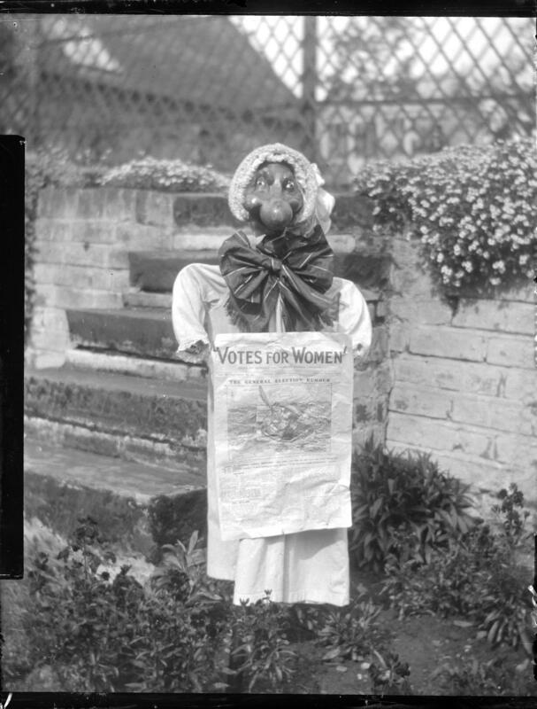 Scarecrow with a bonnet and a big bow, standing in front of a brick wall ,holding a copy of a 'Votes for Women' poster.