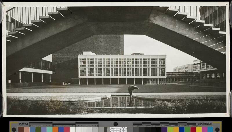 View of a modernist glass building in the medium distance, framed by an elevated concrete pedestrian bridge.